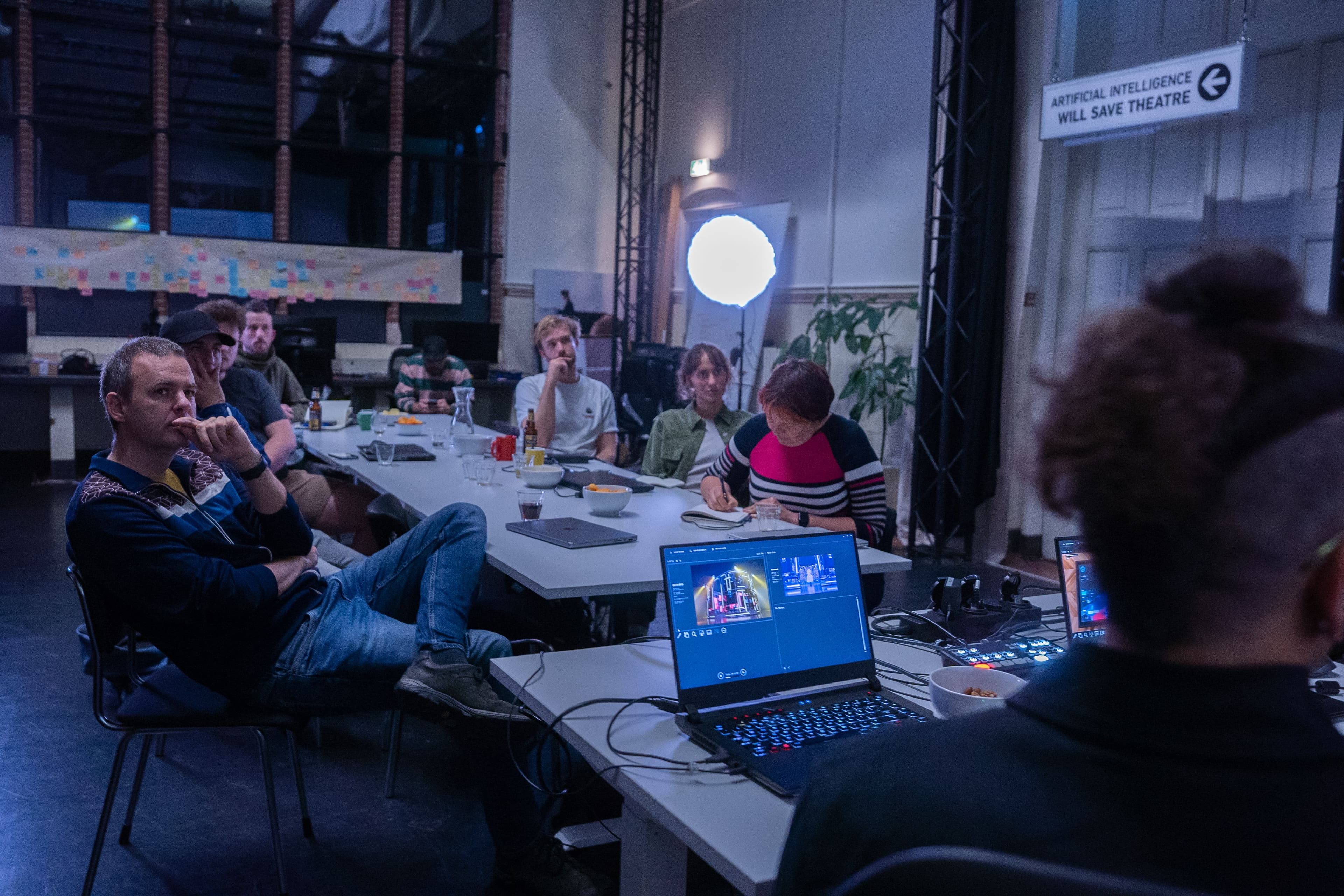 Een groep mensen zit aan een tafel en luistert geconcentreert naar iemand. De spreker zit achter een laptop waarop creative coding software te zien is. 