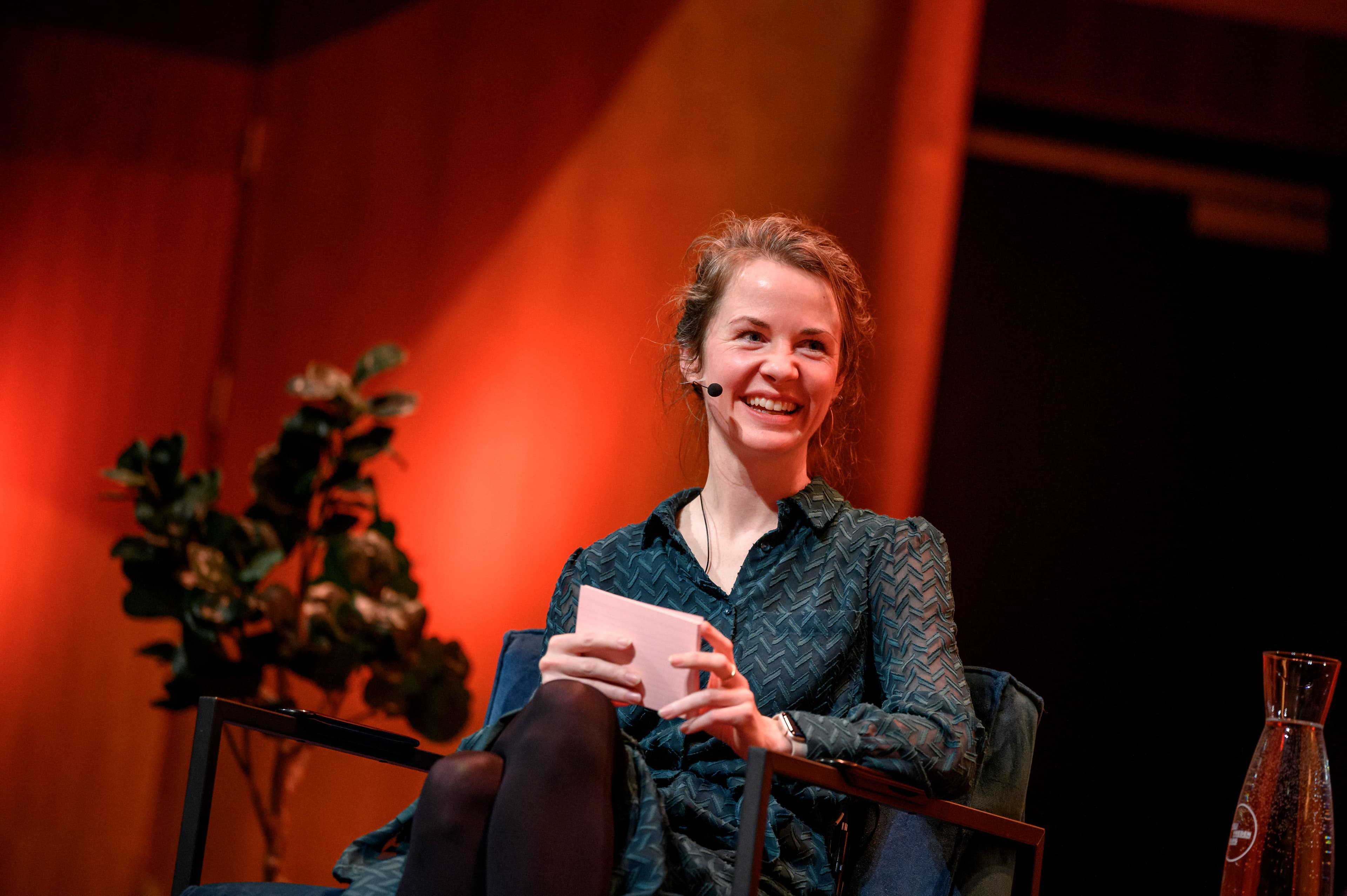 Foto van Anne Breure in een stoel, lachend, met kaartjes in haar hand en een microfoon