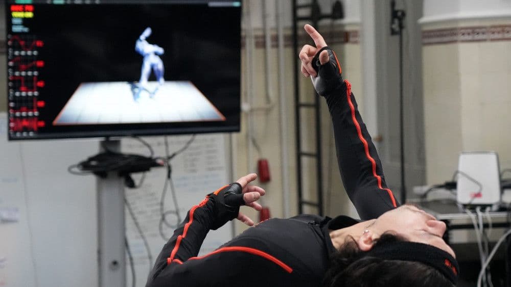 Danser in een motion-capture pak, en een scherm met zijn avatar op de achtergrond.