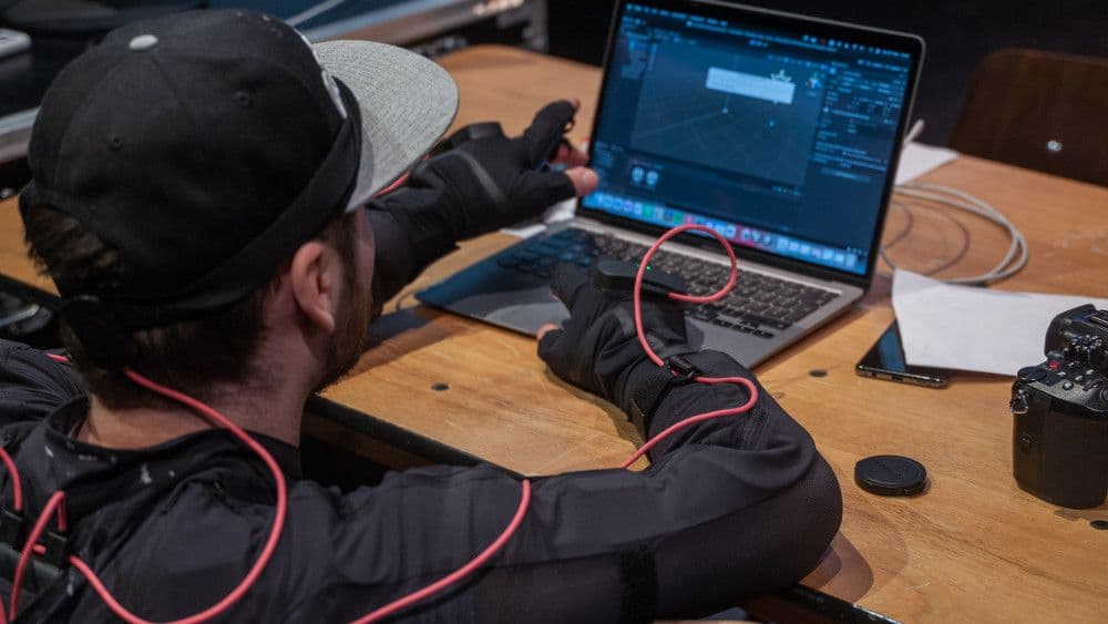  Een persoon in een motion capture-pak werkt aan een laptop met 3D-software, omringd door technische apparatuur op een houten tafel.